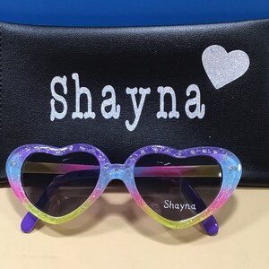 May include: Heart-shaped sunglasses with a rainbow gradient and glitter accents. The sunglasses have a purple frame and the words "Shayna" are printed on the right lens. The sunglasses are in a black case with the name "Shayna" and a silver glitter heart.