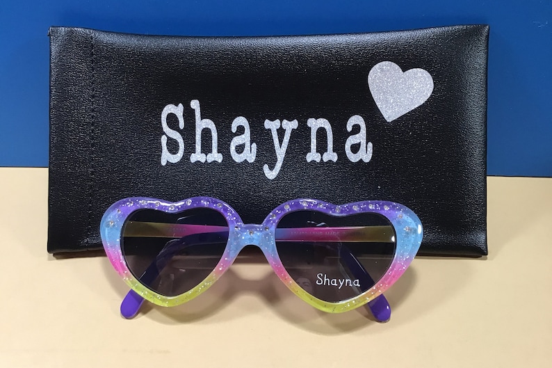 May include: Heart-shaped sunglasses with a rainbow gradient and glitter accents. The sunglasses have a purple frame and the words "Shayna" are printed on the right lens. The sunglasses are in a black case with the name "Shayna" and a silver glitter heart.