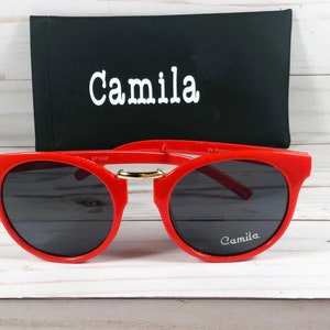 May include: Red round sunglasses with black lenses and a gold accent on the bridge. The sunglasses have the word "Camila" printed on the right lens and the case has the same word printed on the front.
