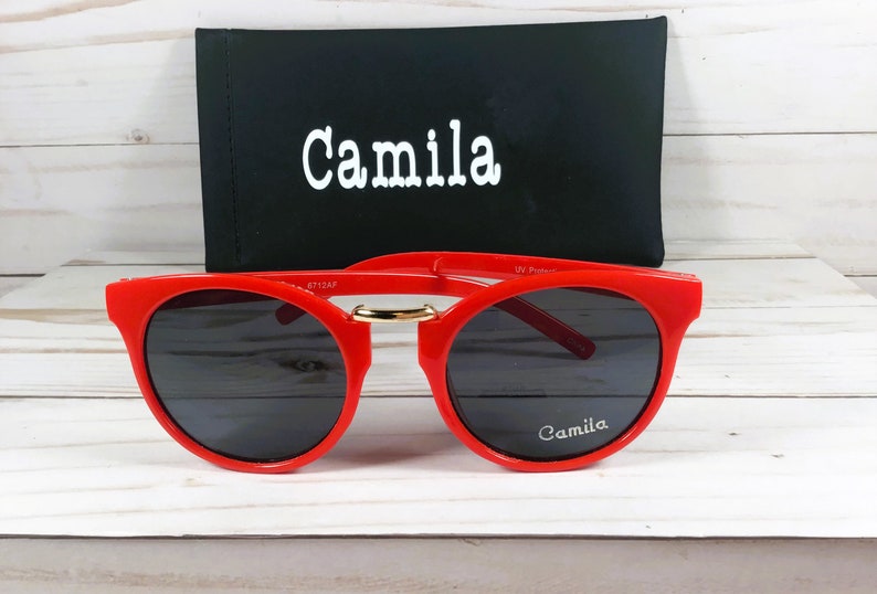 May include: Red round sunglasses with black lenses and a gold accent on the bridge. The sunglasses have the word "Camila" printed on the right lens and the case has the same word printed on the front.