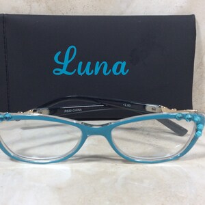 May include: Teal-colored reading glasses with a cat-eye frame, adorned with turquoise-colored embellishments. The glasses rest in front of a black case with the word "Luna" in teal script. The glasses are marked with "+2.00" and "R622 CHINA".