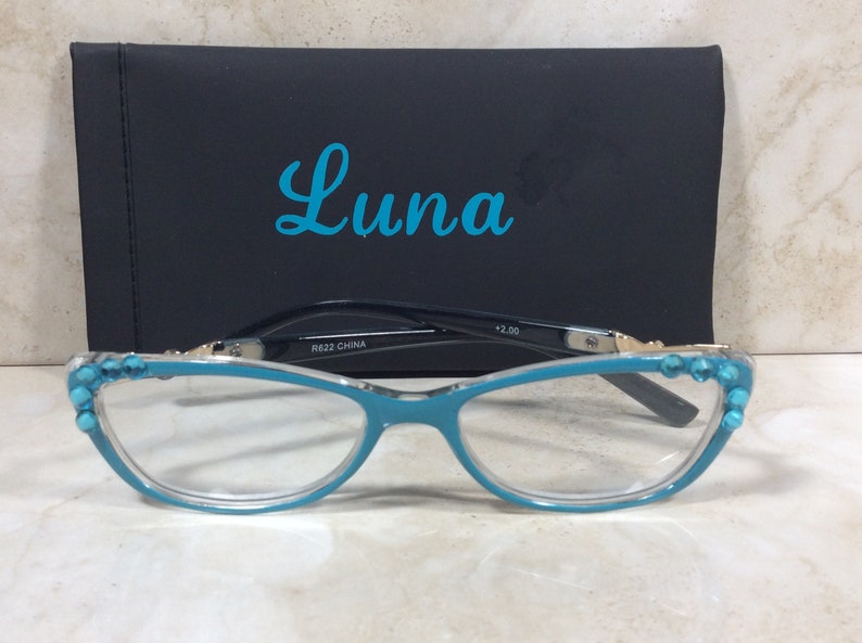 May include: Teal-colored reading glasses with a cat-eye frame, adorned with turquoise-colored embellishments. The glasses rest in front of a black case with the word "Luna" in teal script. The glasses are marked with "+2.00" and "R622 CHINA".