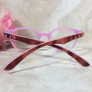 May include: Pink eyeglasses with a cat-eye frame design. The frames are a light pink color with brown temples. Small, clear rhinestones are set into the temples. The glasses are set against a marble background.