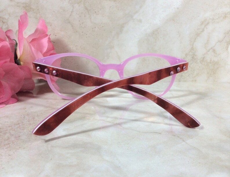 May include: Pink eyeglasses with a cat-eye frame design. The frames are a light pink color with brown temples. Small, clear rhinestones are set into the temples. The glasses are set against a marble background.