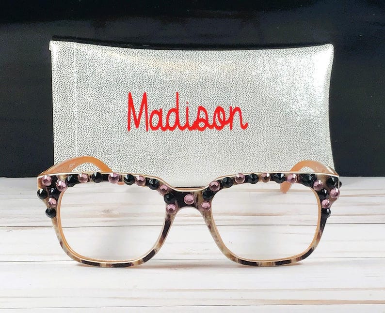 Op de afbeelding: Een bril met een luipaardprint montuur, versierd met zwarte en roze edelstenen. De bril staat voor een zilveren pailletten etui met de naam "Madison" in het rood geborduurd.