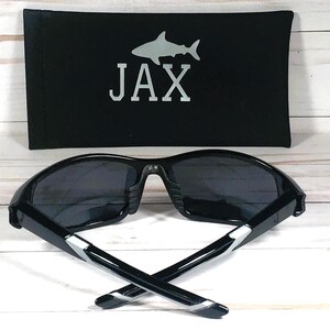 Może przedstawiać: Czarne okulary przeciwsłoneczne z srebrną obw&oacute;dką i sylwetką rekina z napisem "JAX" na czarnym etui.