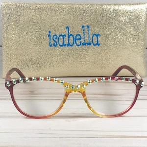 Puede incluir: Gafas con montura degradada en rojo y amarillo que presentan una hilera de pedrer&iacute;a iridiscente en el borde superior. Detr&aacute;s de las gafas hay un estuche rectangular de purpurina dorada con el nombre isabella impreso en letras azules.
