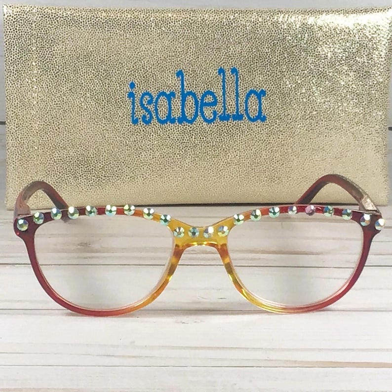 Puede incluir: Gafas con montura degradada en rojo y amarillo que presentan una hilera de pedrer&iacute;a iridiscente en el borde superior. Detr&aacute;s de las gafas hay un estuche rectangular de purpurina dorada con el nombre isabella impreso en letras azules.