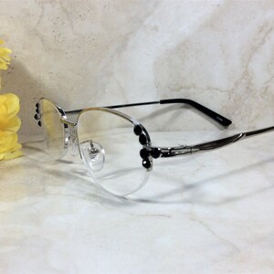 May include: Silver-framed eyeglasses with clear lenses and black accents. The frames feature decorative black embellishments on the sides. The temples are thin and silver, transitioning to black at the ear pieces. A yellow flower is in the background.