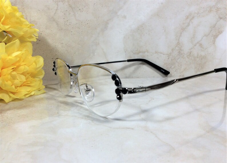 May include: Silver-framed eyeglasses with clear lenses and black accents. The frames feature decorative black embellishments on the sides. The temples are thin and silver, transitioning to black at the ear pieces. A yellow flower is in the background.