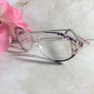 Puede incluir: Gafas transparentes con forma de ojo de gato, con montura plateada y detalles morados. Las patillas est&aacute;n adornadas con peque&ntilde;as gemas rosas. Una flor rosa se encuentra en el fondo.