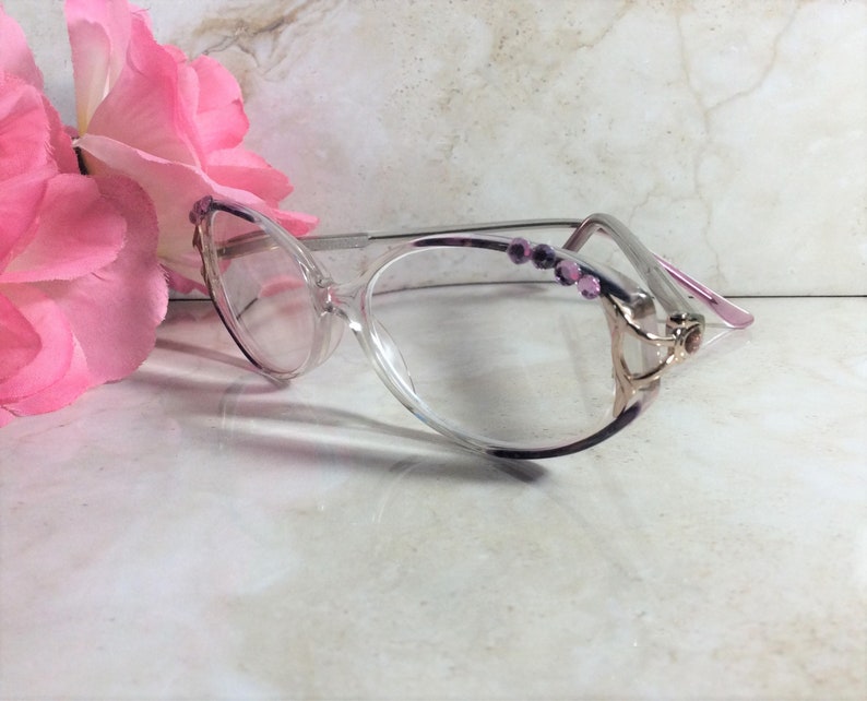 Puede incluir: Gafas transparentes con forma de ojo de gato, con montura plateada y detalles morados. Las patillas est&aacute;n adornadas con peque&ntilde;as gemas rosas. Una flor rosa se encuentra en el fondo.