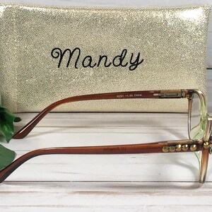 Pu&ograve; includere: Occhiali con montatura marrone e abbellimenti dorati davanti a una pochette dorata con paillettes. La pochette ha il nome "Mandy" ricamato in corsivo nero. Gli occhiali hanno una forma rettangolare e sono su una superficie di legno bianco.