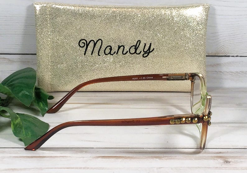Pu&ograve; includere: Occhiali con montatura marrone e abbellimenti dorati davanti a una pochette dorata con paillettes. La pochette ha il nome "Mandy" ricamato in corsivo nero. Gli occhiali hanno una forma rettangolare e sono su una superficie di legno bianco.