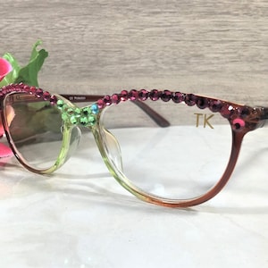 May include: Eyeglasses with a gradient green and brown frame, adorned with pink and green rhinestones. The top of the frame is embellished with a row of pink rhinestones. The initials "TK" are visible on the right temple.