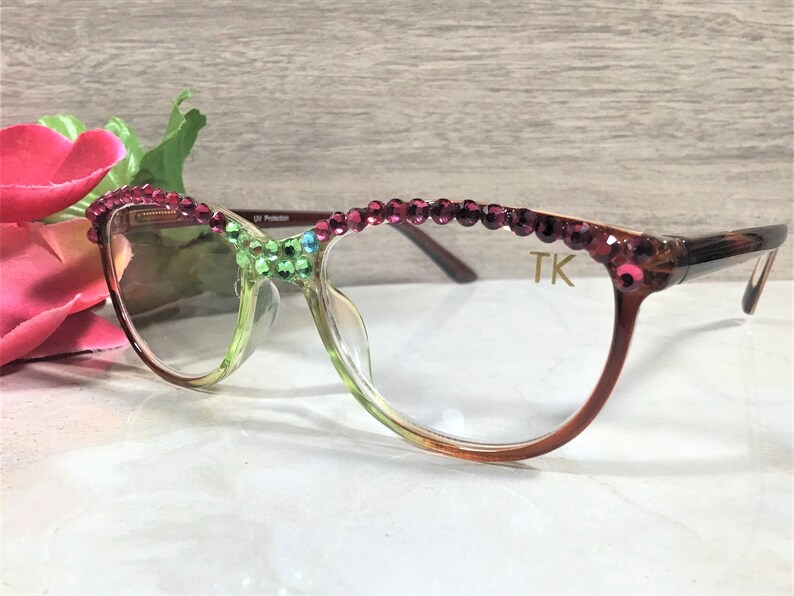 May include: Eyeglasses with a gradient green and brown frame, adorned with pink and green rhinestones. The top of the frame is embellished with a row of pink rhinestones. The initials "TK" are visible on the right temple.