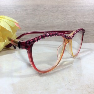 May include: Reading glasses with a gradient red and yellow frame, adorned with sparkling magenta rhinestones along the top. The glasses are set against a soft background with a yellow and pink flower.