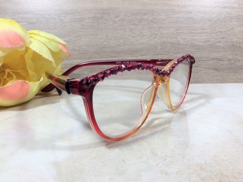 May include: Reading glasses with a gradient red and yellow frame, adorned with sparkling magenta rhinestones along the top. The glasses are set against a soft background with a yellow and pink flower.