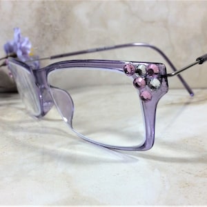 May include: Light purple translucent eyeglasses featuring a cluster of pink and clear rhinestones on the outer top corners of the frames. The glasses have thin, metallic silver-toned temples and clear lenses, showcasing a decorative embellishment.