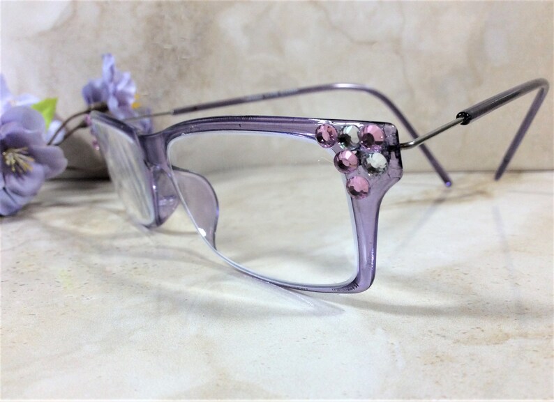 May include: Light purple translucent eyeglasses featuring a cluster of pink and clear rhinestones on the outer top corners of the frames. The glasses have thin, metallic silver-toned temples and clear lenses, showcasing a decorative embellishment.