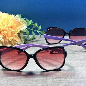 May include: Two pairs of sunglasses with black frames and purple arms. The lenses have a gradient from red to purple. A yellow and white flower arrangement is in the background.