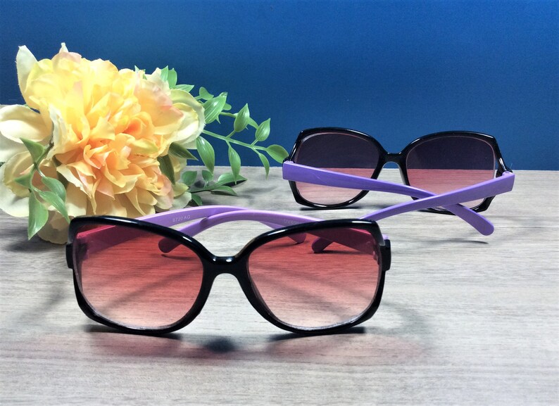 May include: Two pairs of sunglasses with black frames and purple arms. The lenses have a gradient from red to purple. A yellow and white flower arrangement is in the background.
