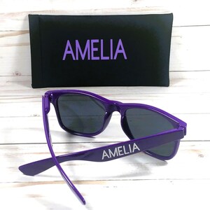 May include: Purple sunglasses with the name Amelia printed on the arm. The sunglasses come with a black case that also has the name Amelia printed on it.