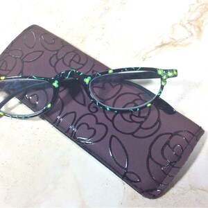 K&ouml;nnte beinhalten: Lesebrille mit schwarzem Rahmen, verziert mit einem Blumenmuster und gr&uuml;nen Juwelen, auf einem dunkelvioletten Etui mit Rosenmuster. Die Brille hat ein t&uuml;rkis-gelbes Blumenmuster.