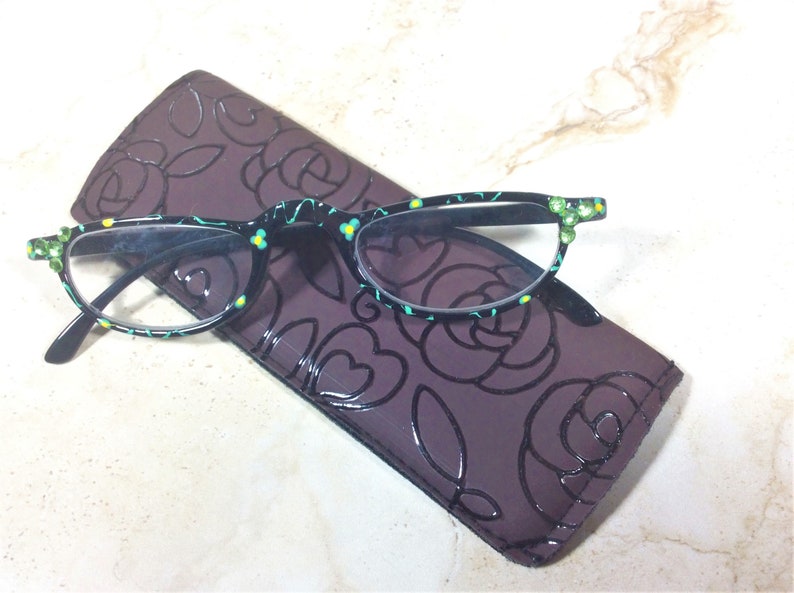 K&ouml;nnte beinhalten: Lesebrille mit schwarzem Rahmen, verziert mit einem Blumenmuster und gr&uuml;nen Juwelen, auf einem dunkelvioletten Etui mit Rosenmuster. Die Brille hat ein t&uuml;rkis-gelbes Blumenmuster.