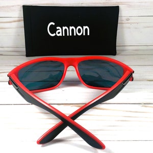 K&ouml;nnte beinhalten: Rote Sonnenbrille mit schwarzen B&uuml;geln und blauen Gl&auml;sern. Die Sonnenbrille ist zusammengeklappt und das Etui ist im Hintergrund sichtbar. Das Etui ist schwarz mit dem in Wei&szlig; gedruckten Wort "Cannon".