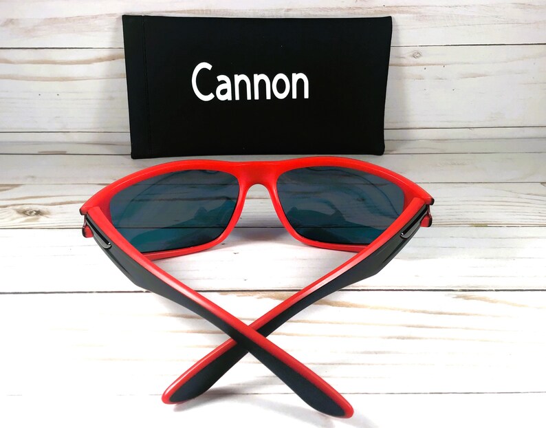 K&ouml;nnte beinhalten: Rote Sonnenbrille mit schwarzen B&uuml;geln und blauen Gl&auml;sern. Die Sonnenbrille ist zusammengeklappt und das Etui ist im Hintergrund sichtbar. Das Etui ist schwarz mit dem in Wei&szlig; gedruckten Wort "Cannon".