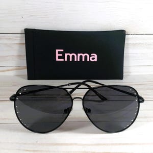 Peut inclure: Lunettes de soleil aviateur noires avec un &eacute;tui noir. Les lunettes ont une monture en m&eacute;tal et des verres fonc&eacute;s. L'&eacute;tui est noir avec le nom "Emma" en lettres roses. Les lunettes ont de petits strass d&eacute;coratifs autour des verres.