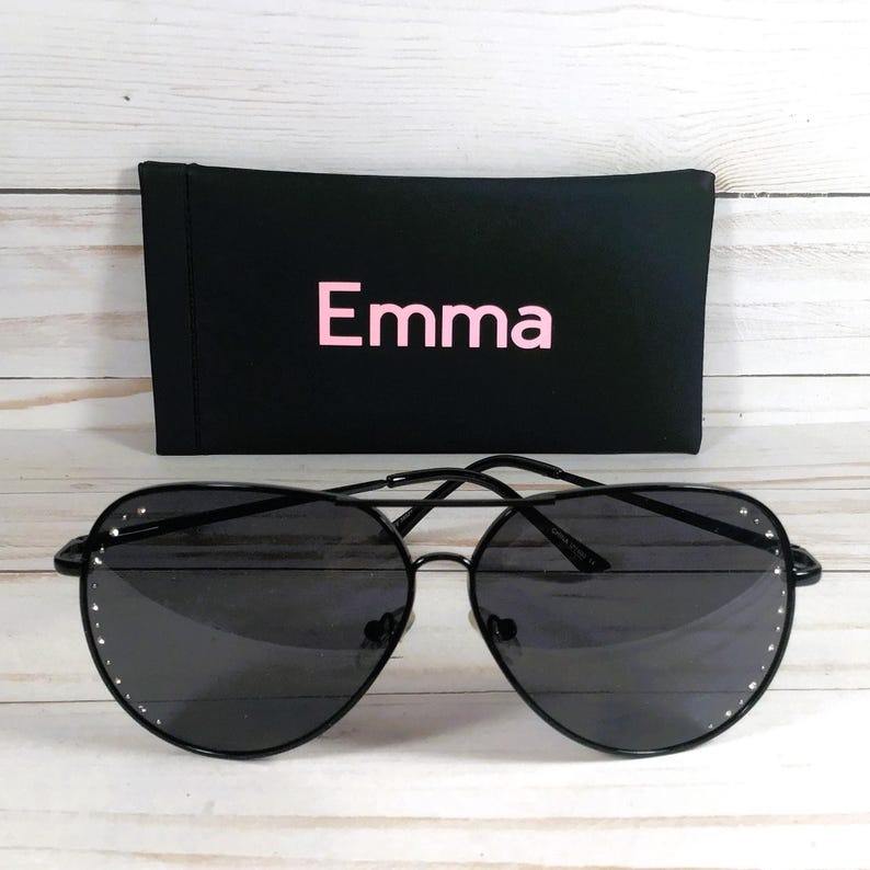 Peut inclure: Lunettes de soleil aviateur noires avec un &eacute;tui noir. Les lunettes ont une monture en m&eacute;tal et des verres fonc&eacute;s. L'&eacute;tui est noir avec le nom "Emma" en lettres roses. Les lunettes ont de petits strass d&eacute;coratifs autour des verres.