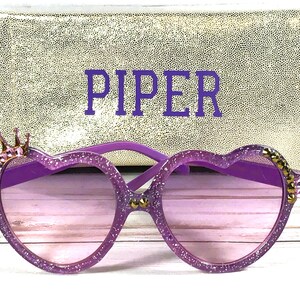 May include: Purple heart-shaped sunglasses with glitter and gold accents, including a crown on the left side. The sunglasses come with a gold glitter case with the name "PIPER" embroidered on it.