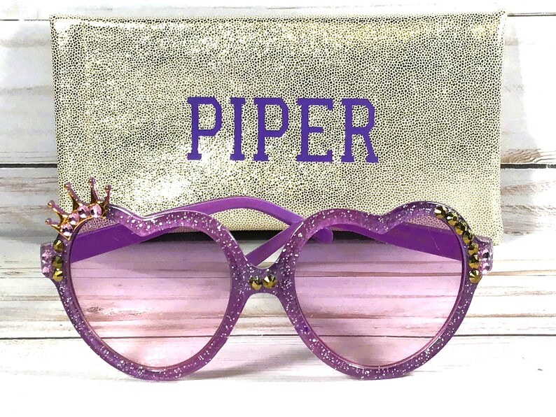 May include: Purple heart-shaped sunglasses with glitter and gold accents, including a crown on the left side. The sunglasses come with a gold glitter case with the name "PIPER" embroidered on it.