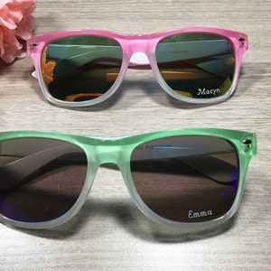 May include: Two pairs of sunglasses with UV protection. The top pair is pink and white with the name "Macyn" on the right lens. The bottom pair is green and white with the name "Emma" on the right lens.