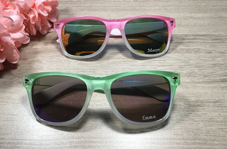 May include: Two pairs of sunglasses with UV protection. The top pair is pink and white with the name "Macyn" on the right lens. The bottom pair is green and white with the name "Emma" on the right lens.