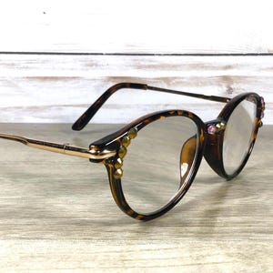May include: Brown tortoiseshell eyeglasses with gold-tone arms and decorative embellishments. The round frames are adorned with small, gold-colored studs and pink gems. The lenses are clear.