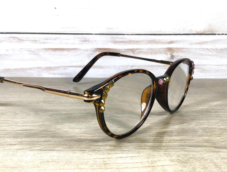 May include: Brown tortoiseshell eyeglasses with gold-tone arms and decorative embellishments. The round frames are adorned with small, gold-colored studs and pink gems. The lenses are clear.