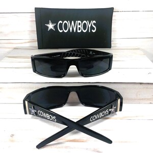 May include: Black sunglasses with the word "COWBOYS" and a star logo on the temples. The sunglasses are displayed with a black case that also has the word "COWBOYS" and a star logo. The sunglasses have a rectangular frame.
