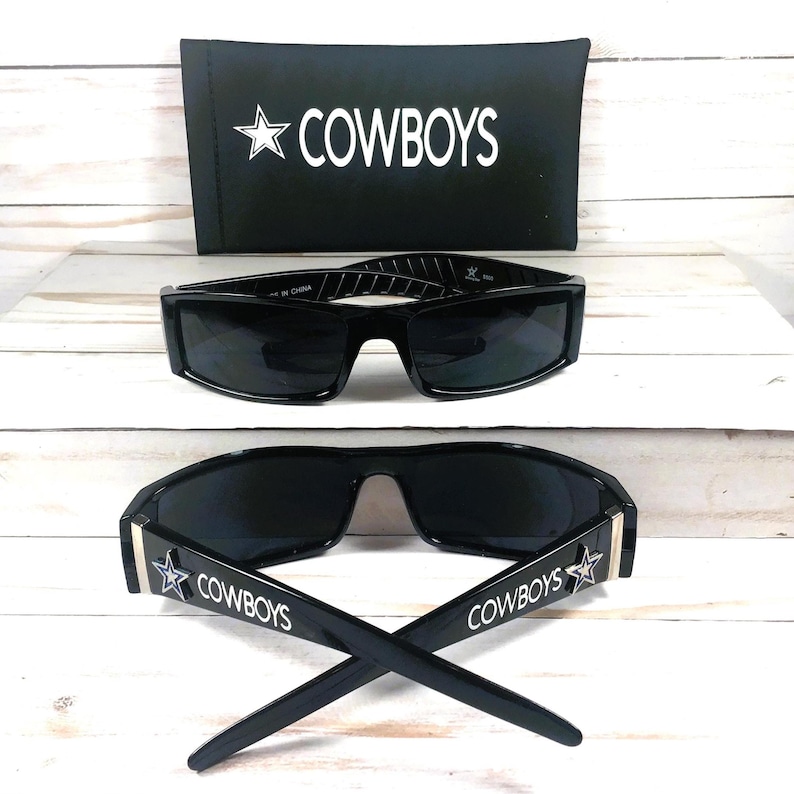 May include: Black sunglasses with the word "COWBOYS" and a star logo on the temples. The sunglasses are displayed with a black case that also has the word "COWBOYS" and a star logo. The sunglasses have a rectangular frame.