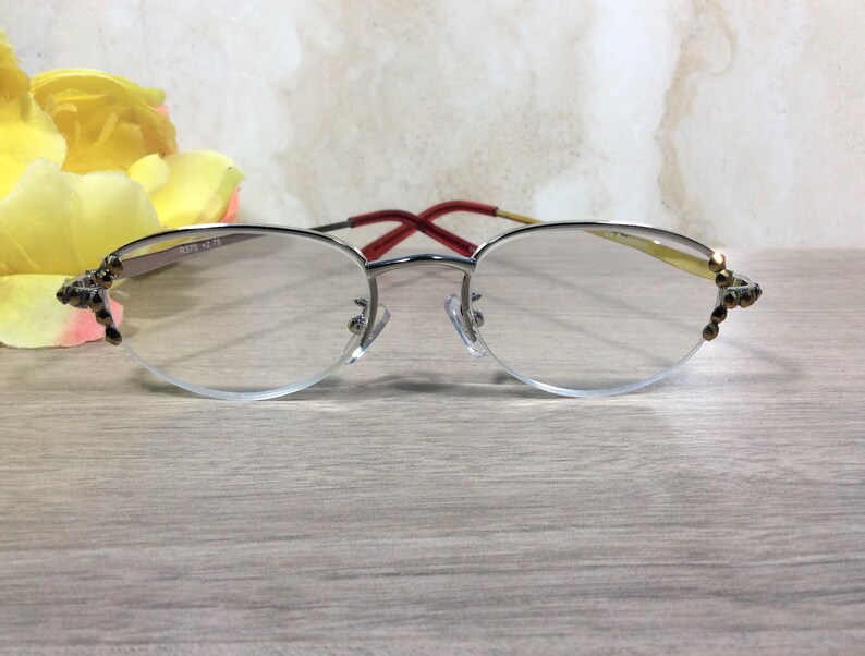 Puede incluir: Gafas de lectura con montura plateada y dise&ntilde;o de media montura. Las gafas tienen una patilla roja y dorada y est&aacute;n adornadas con detalles decorativos en los bordes exteriores. Las lentes son transparentes y la montura incluye el texto "8375 +2.75".