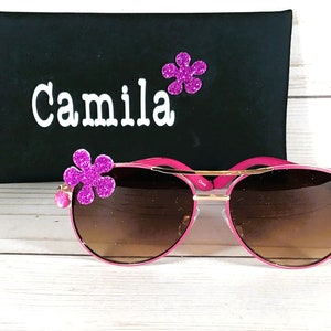Pu&ograve; includere: Occhiali da sole aviator rosa con lenti marroni e dettagli dorati. Gli occhiali hanno un abbellimento a forma di fiore rosa su ogni lato. Gli occhiali sono in una custodia nera con il nome "Camila" scritto su di essa.