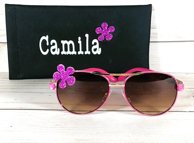 Pu&ograve; includere: Occhiali da sole aviator rosa con lenti marroni e dettagli dorati. Gli occhiali hanno un abbellimento a forma di fiore rosa su ogni lato. Gli occhiali sono in una custodia nera con il nome "Camila" scritto su di essa.