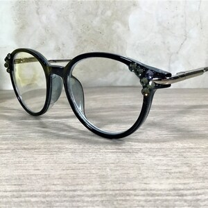 K&ouml;nnte beinhalten: Schwarze Brille mit klaren Gl&auml;sern und d&uuml;nnen Metallb&uuml;geln. Die &auml;u&szlig;eren Ecken der schwarzen Fassung sind mit kleinen, dunklen, facettierten Ziersteinen besetzt, die das Licht einfangen und dem Brillendesign ein dezentes Funkeln verleihen.