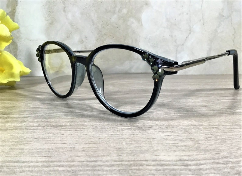 K&ouml;nnte beinhalten: Schwarze Brille mit klaren Gl&auml;sern und d&uuml;nnen Metallb&uuml;geln. Die &auml;u&szlig;eren Ecken der schwarzen Fassung sind mit kleinen, dunklen, facettierten Ziersteinen besetzt, die das Licht einfangen und dem Brillendesign ein dezentes Funkeln verleihen.