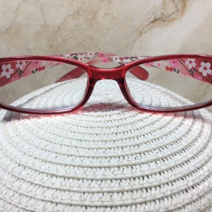Puede incluir: Gafas con montura roja con un estampado floral en las patillas y pedrer&iacute;a decorativa en las esquinas. Las lentes transparentes descansan sobre una superficie blanca texturizada. El dise&ntilde;o es femenino y elegante.