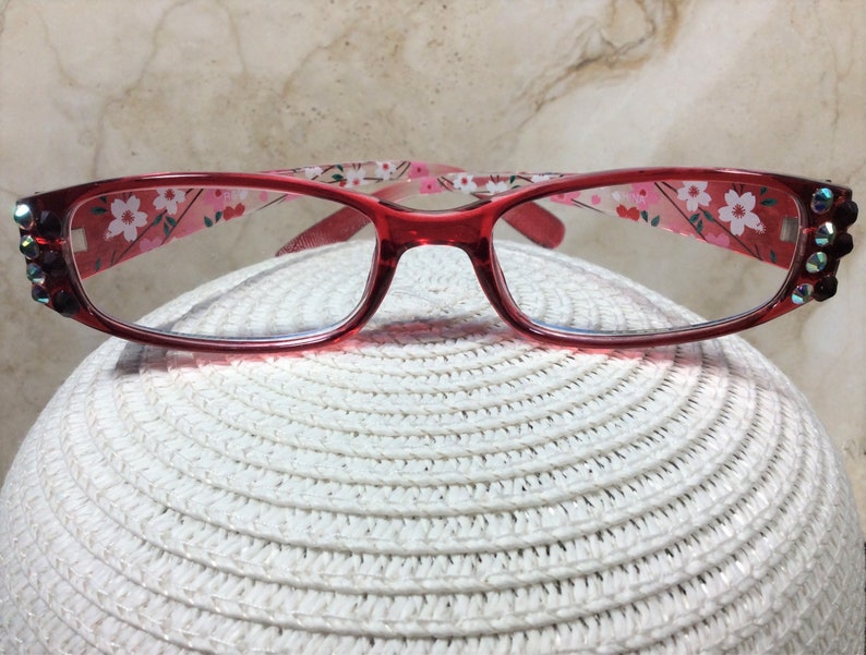 Puede incluir: Gafas con montura roja con un estampado floral en las patillas y pedrer&iacute;a decorativa en las esquinas. Las lentes transparentes descansan sobre una superficie blanca texturizada. El dise&ntilde;o es femenino y elegante.