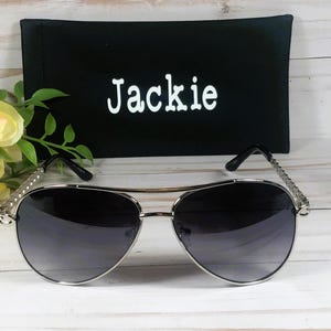 Peut inclure: Lunettes de soleil aviateur argent&eacute;es avec des verres gris fonc&eacute; et un &eacute;tui en tissu noir avec le nom "Jackie" brod&eacute; dessus.