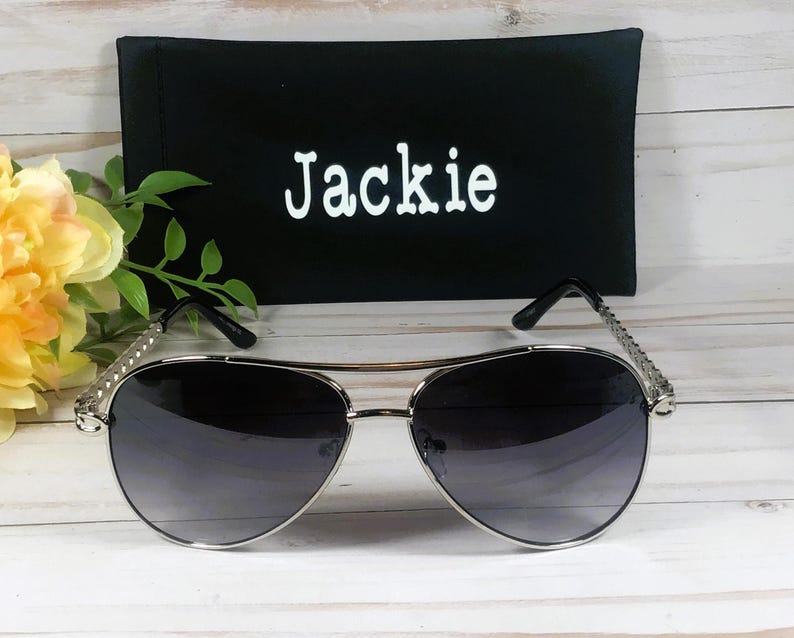 Peut inclure: Lunettes de soleil aviateur argent&eacute;es avec des verres gris fonc&eacute; et un &eacute;tui en tissu noir avec le nom "Jackie" brod&eacute; dessus.